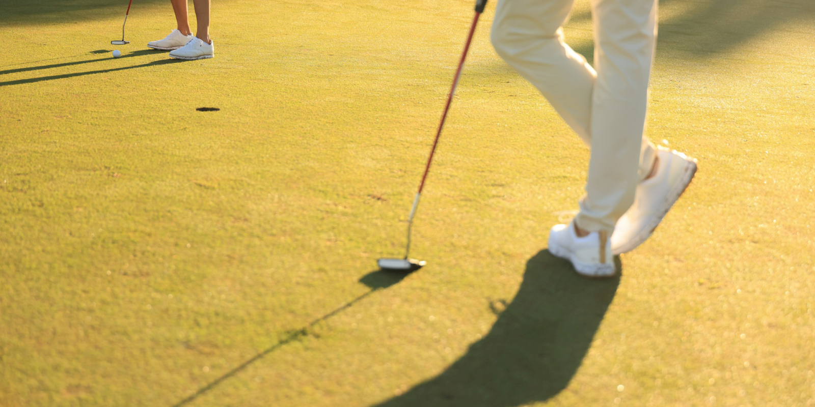 Two golfers on a green: one putter near the ball while a second player watches, shadows long on the fairway under late sunlight.