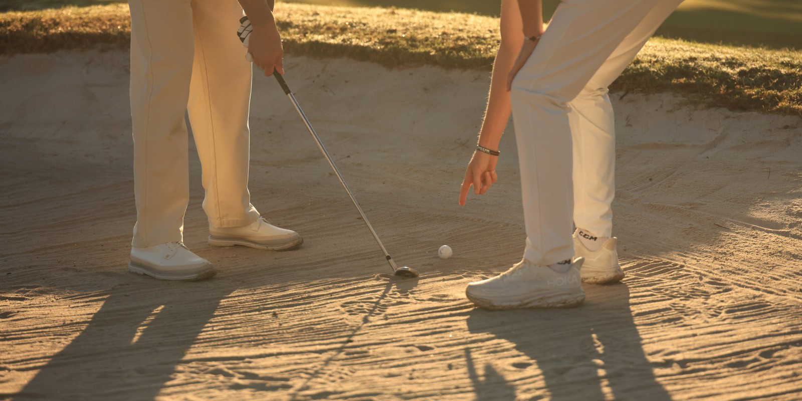 Two golfers practice a chip shot on a sand bunker, low sun casting long shadows as the ball nears the hole.