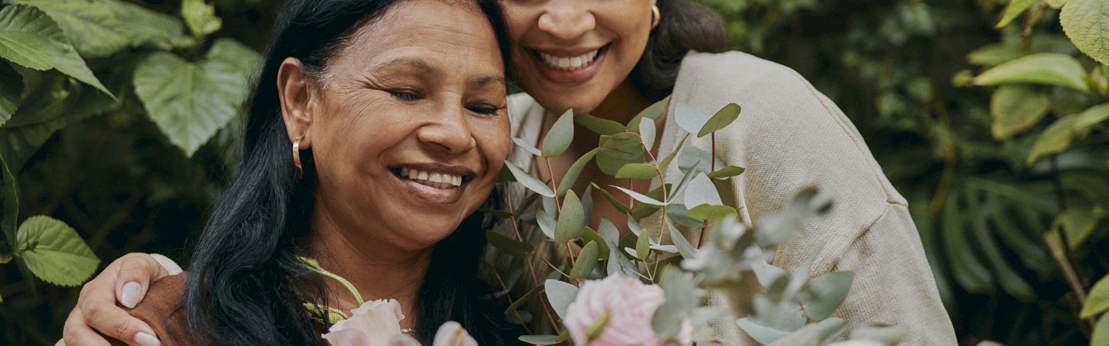 Two people are embracing in a garden setting, smiling and holding a bouquet of flowers.
