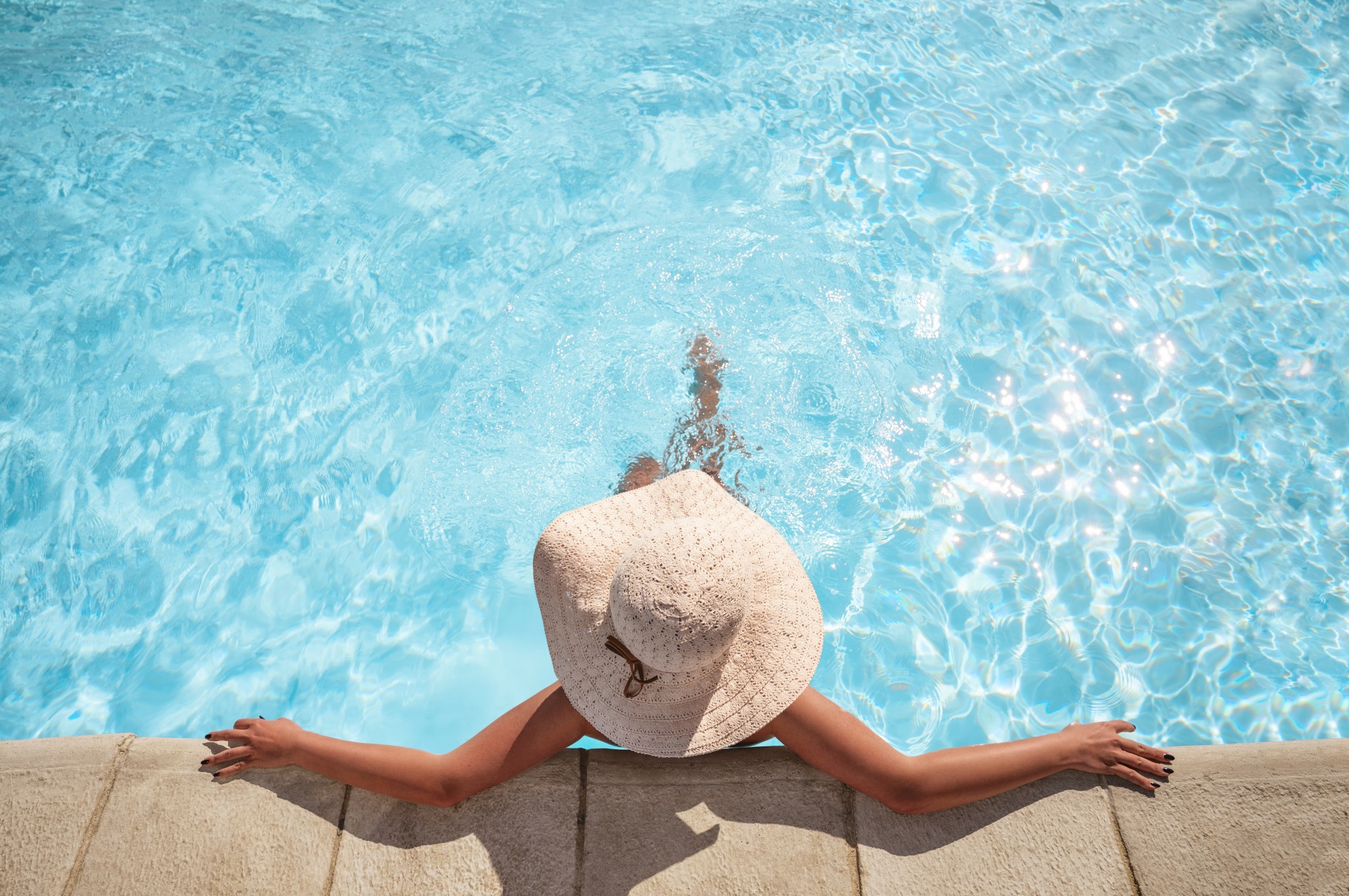 A person wearing a sun hat relaxes in a pool, sitting at the edge with the legs submerged in clear blue water.