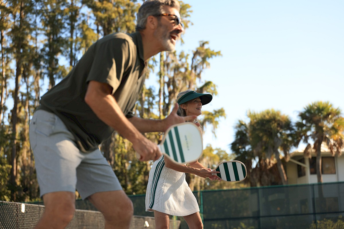 An older man and woman are playing pickleball outdoors on a court with trees in the background.