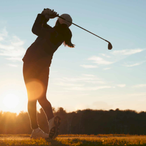 A golfer is swinging a club on a course at sunset, with the sun casting a warm glow in the background.