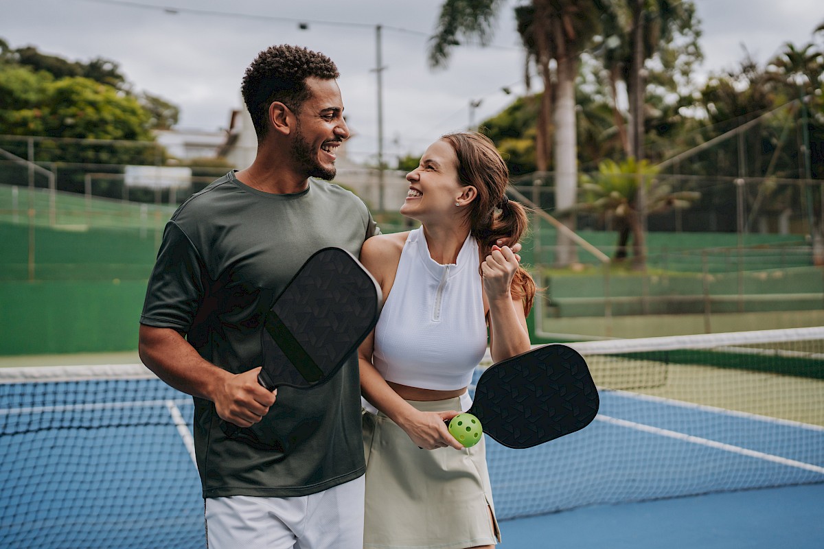 Two people stand on a pickleball court, smiling while holding paddles and a ball.