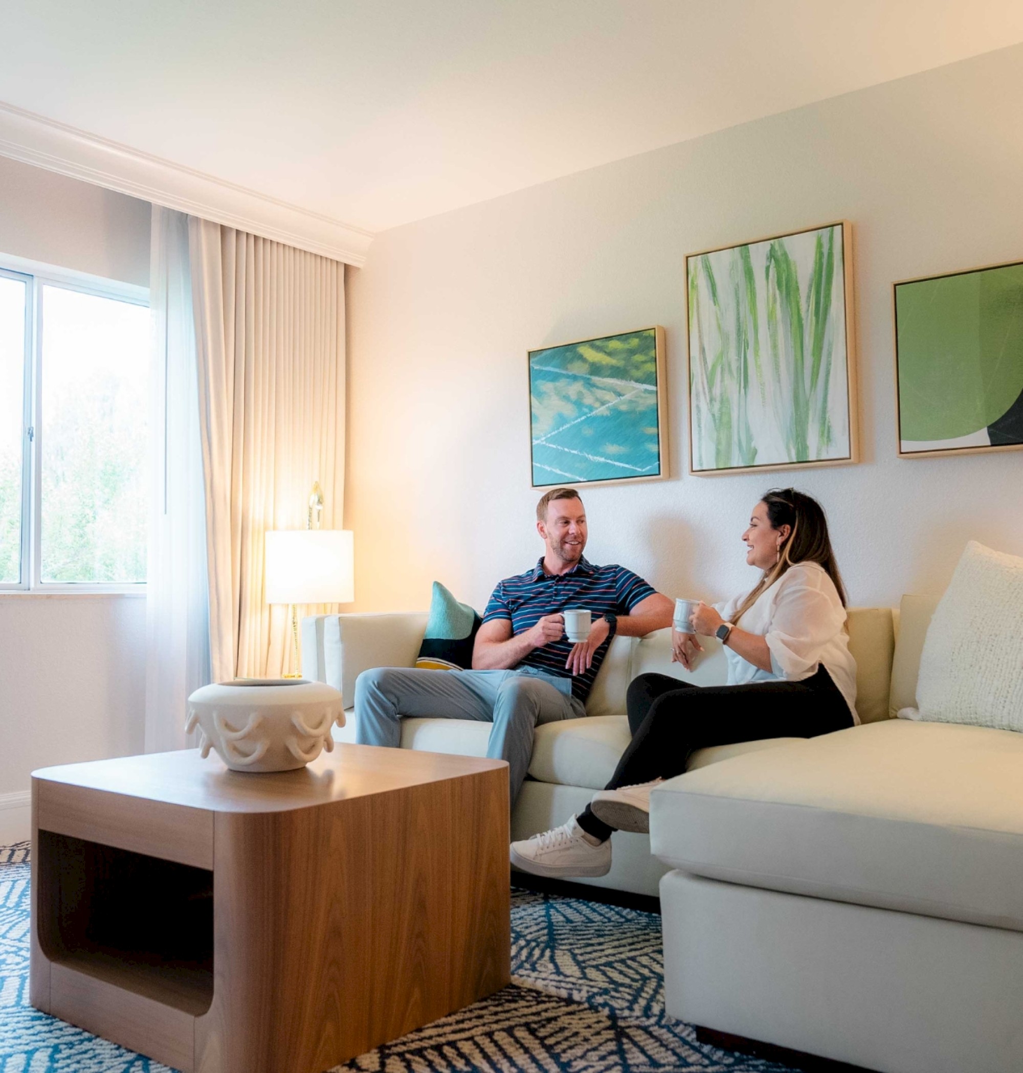 A cozy living room with two people sitting on a sofa, chatting. Artwork hangs on the wall, and natural light streams through the window.