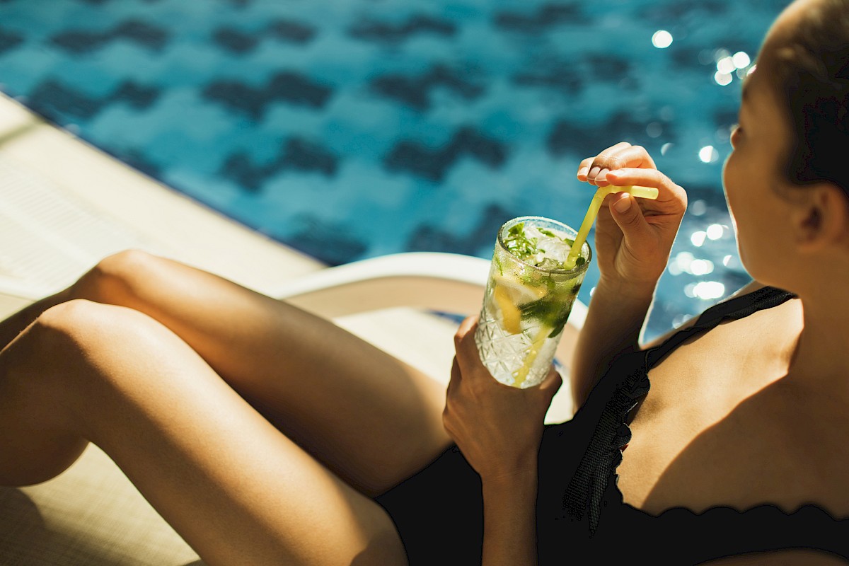 A person in a black swimsuit relaxes by a pool, holding a refreshing drink with mint and a straw, enjoying a sunny day.