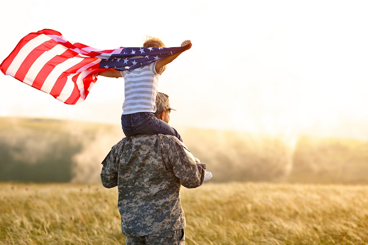 A child on a person's shoulders, draped with an American flag, standing in a field at sunset or sunrise.
