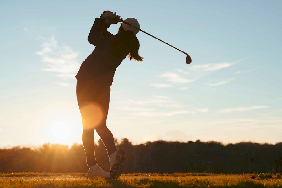 A person is playing golf at sunrise or sunset, finishing a swing. The sun is low on the horizon, creating a silhouette effect.