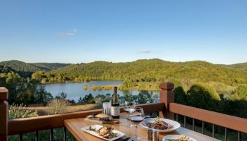 A scenic lakeside view from a balcony with a table set for two, featuring gourmet dishes, wine, and glasses against a backdrop of hills and trees.