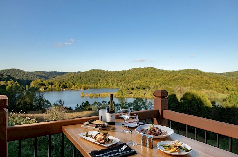 A scenic lakeside view from a balcony with a table set for two, featuring gourmet dishes, wine, and glasses against a backdrop of hills and trees.