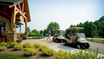 A person is walking towards a golf course clubhouse with golf carts parked nearby on a sunny day, surrounded by greenery and flowers.