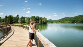 Two people stand on a wooden boardwalk overlooking a tranquil lake surrounded by green hills and trees under a blue sky with a few clouds.