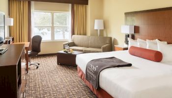 A well-furnished hotel room with a bed, sofa, desk, chair, TV, and window with yellow curtains, featuring modern decor and amenities.