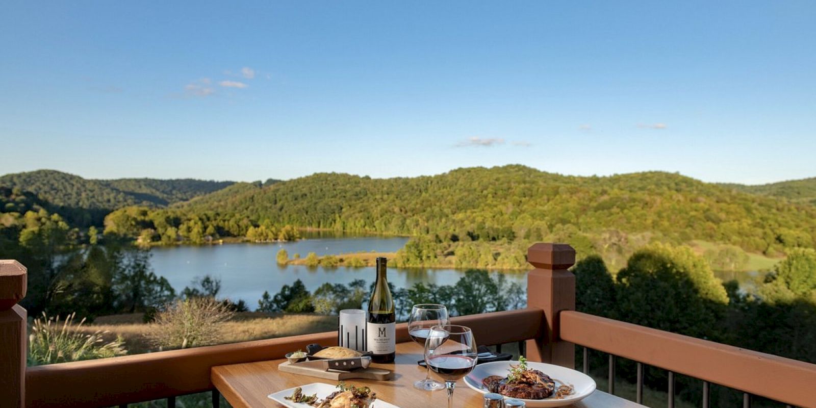 A table with two glasses of wine and gourmet meals is set on a balcony overlooking a serene lake and lush green hills under a clear blue sky.