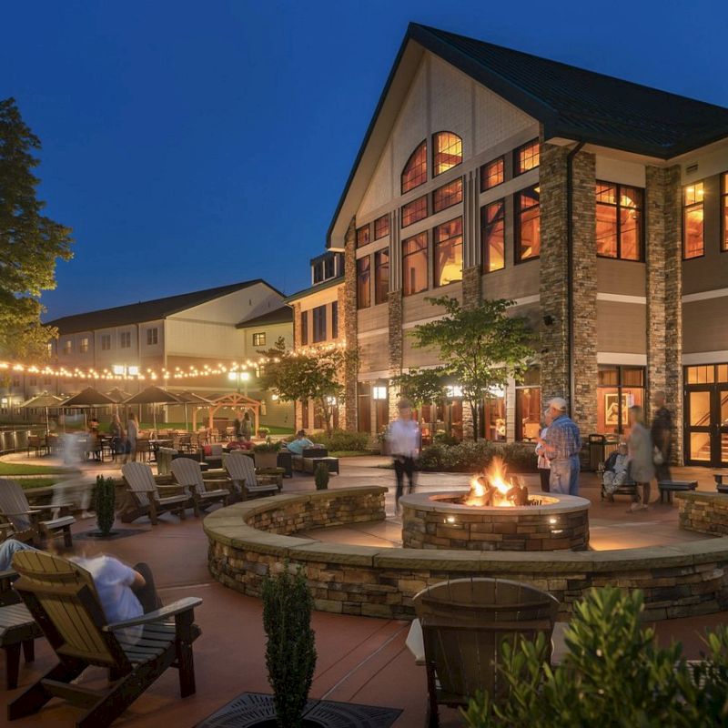 An outdoor seating area with a fire pit, lounge chairs, string lights, and people enjoying the evening near a large, well-lit building.