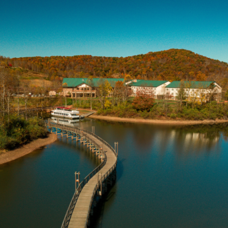 A serene lake with a curved pier leading to buildings with green roofs, surrounded by autumn trees and reflecting water under a clear blue sky.