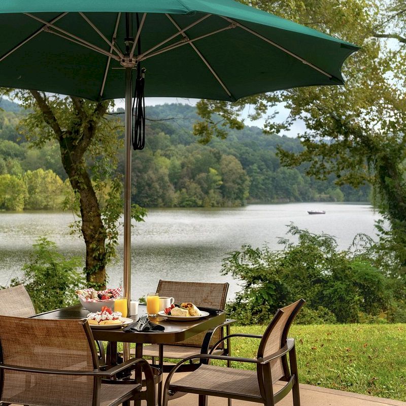 An outdoor table with four chairs and an umbrella, set with breakfast items, overlooking a serene lake and lush green landscape.