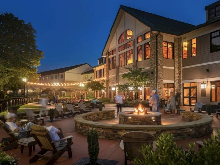 An evening scene of a well-lit building with a fire pit surrounded by chairs on a patio, people around, and string lights decorating the area.