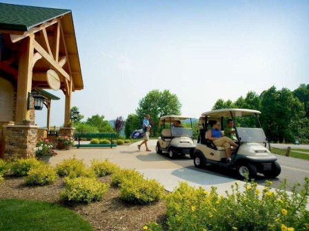 The image shows people near a building with wooden accents and a green roof, featuring two golf carts on a path surrounded by greenery.