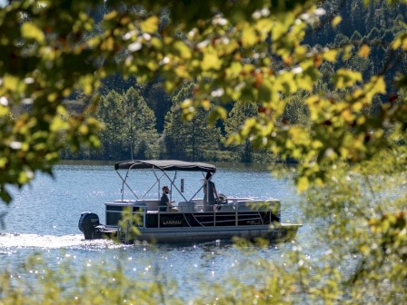 A boat with people on a lake, surrounded by trees and mountains in the background, framed by leaves in the foreground. The water looks calm and serene.