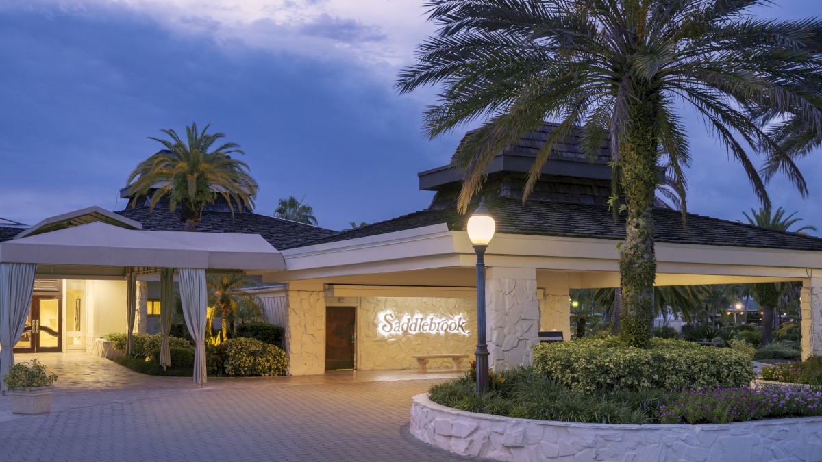 A building entrance surrounded by palm trees and greenery, featuring a lit sign that reads 