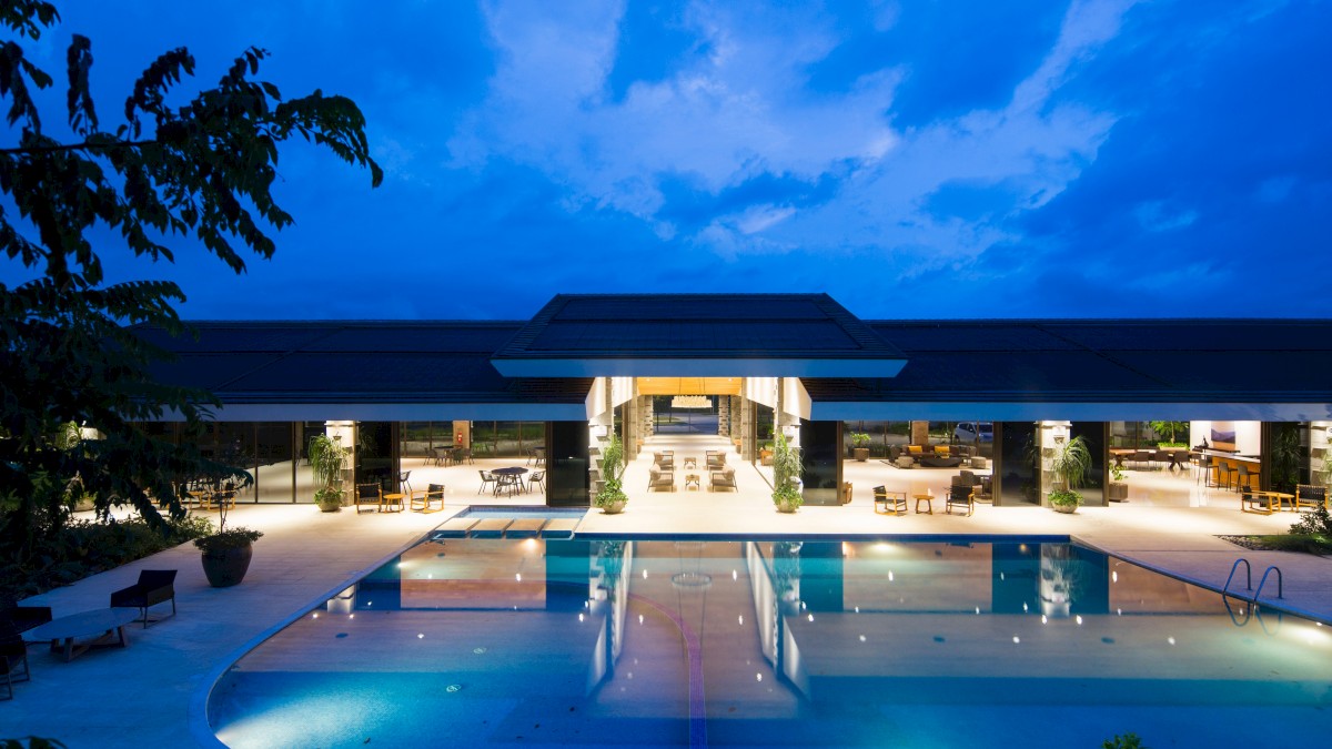 A luxurious building with an open-area design, featuring a large, lit swimming pool in the foreground and outdoor seating areas.