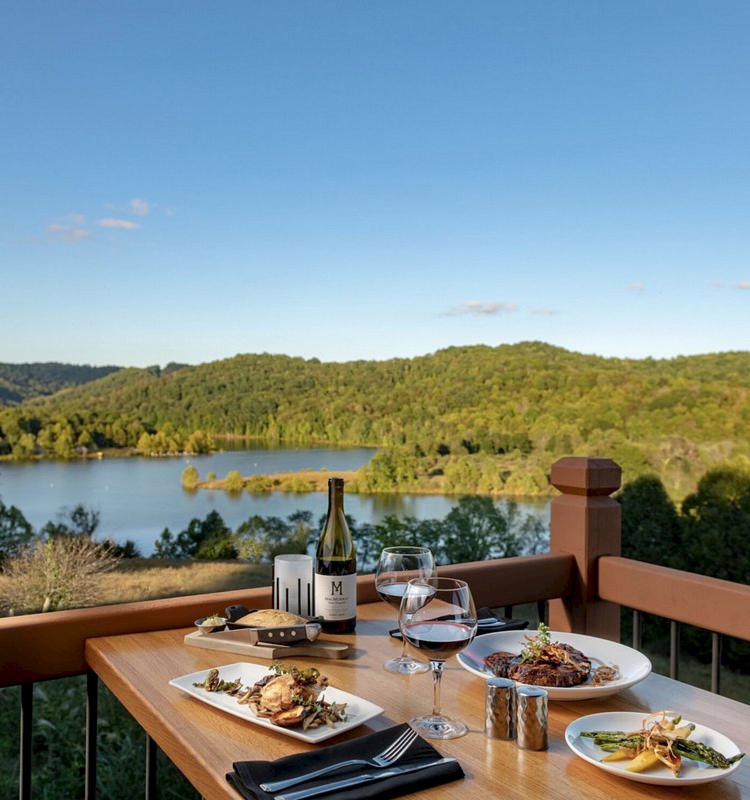 A table set for a meal with wine, dishes, and utensils on a balcony overlooking a scenic lake and forested hills under a clear blue sky.