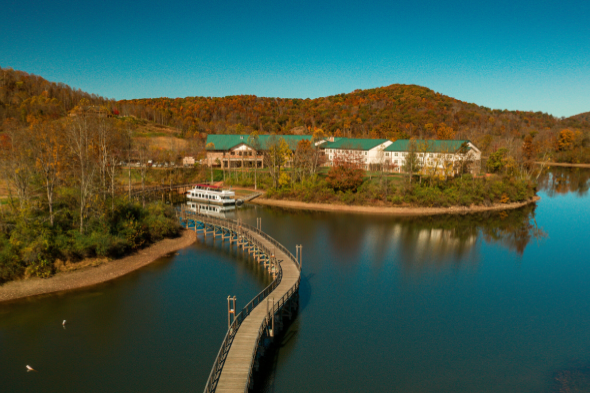 A scenic view of a lake with a winding dock, buildings surrounded by trees with fall foliage, and hills in the background under a clear sky.