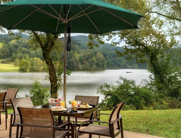 An outdoor dining area with a table and chairs under a green umbrella, set with breakfast items, overlooks a scenic lake and wooded landscape.