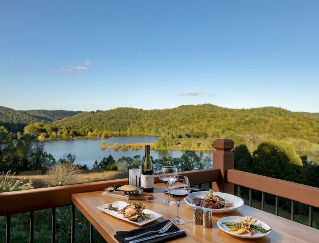 A scenic outdoor dining setup overlooking a lake and green hills, with two plates of food, wine, and glasses on a wooden table.