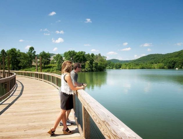 Two people are standing on a wooden pathway by a lake, surrounded by greenery and hills under a clear blue sky.