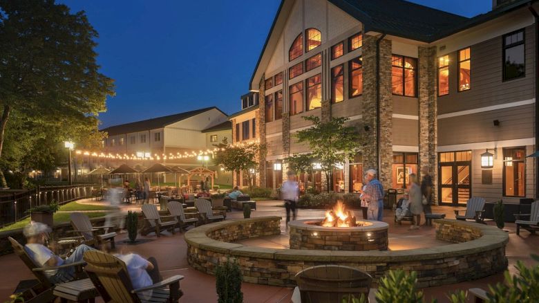 People gather around an outdoor fire pit at night by a large building, with string lights overhead and patio seating surrounding the area.