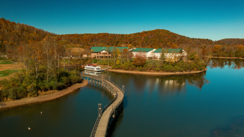 A serene lake with clear blue water, a curving walkway leading to a docked boat, and buildings with green roofs set against a backdrop of autumn trees.