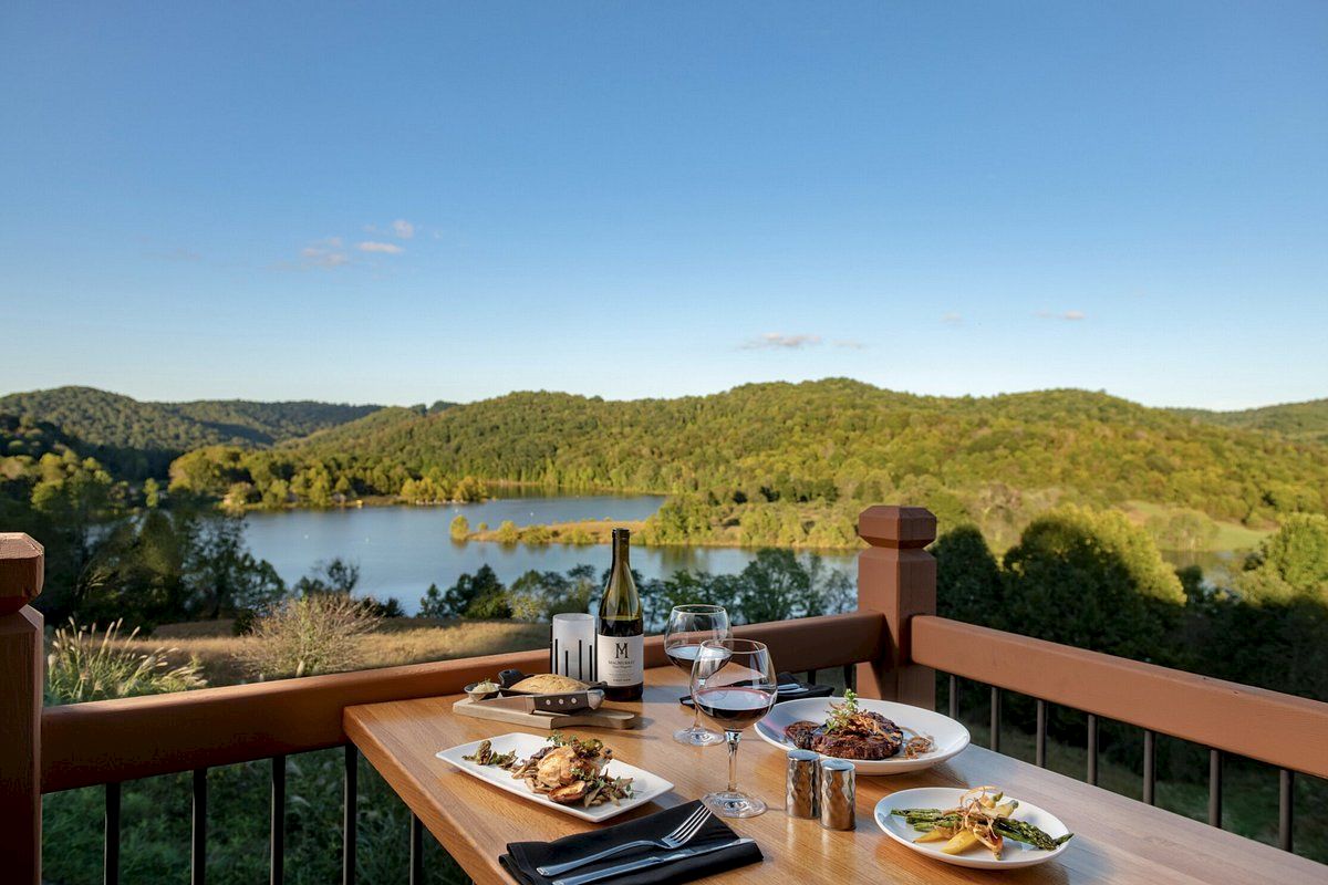 Dinner table overlooking a scenic lake and green hills. Plates of food, wine bottle, and glasses are on the table. Clear sky with few clouds.