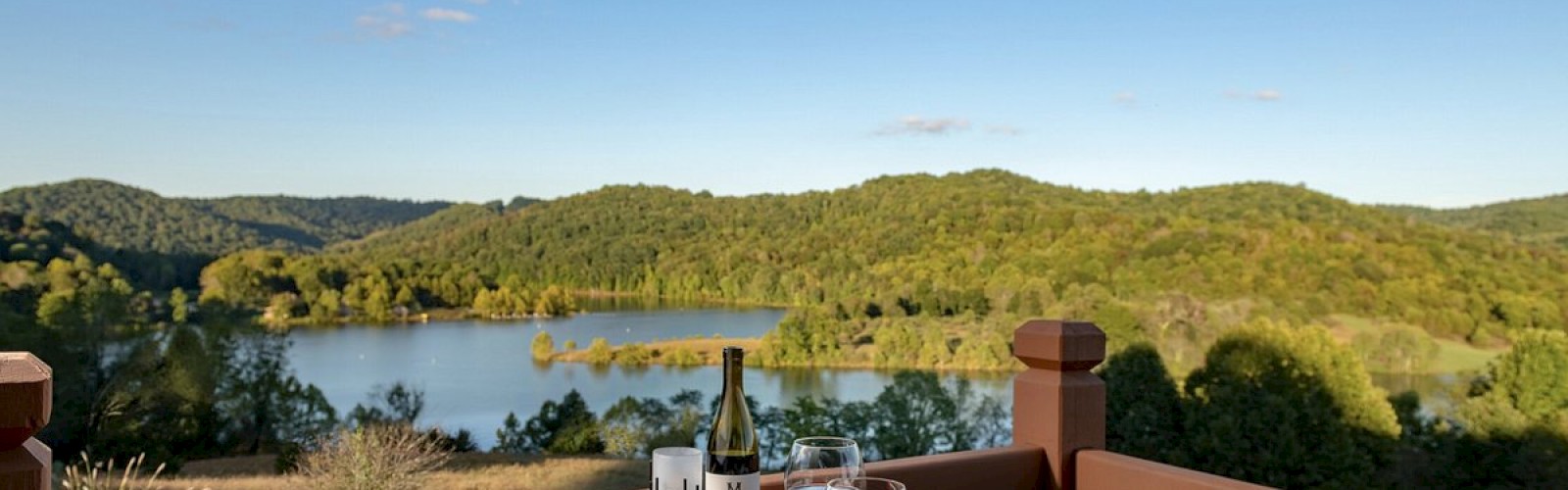 A scenic outdoor dining setup on a wooden table features meals, glasses of wine, and a bottle, overlooking a serene lake and green hills under a clear sky.