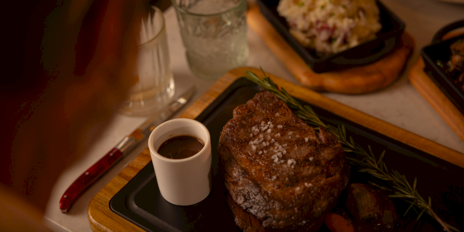 A steak with coarse salt, a cup of sauce, and rosemary on a wooden board. A side dish and drinks are in the background.