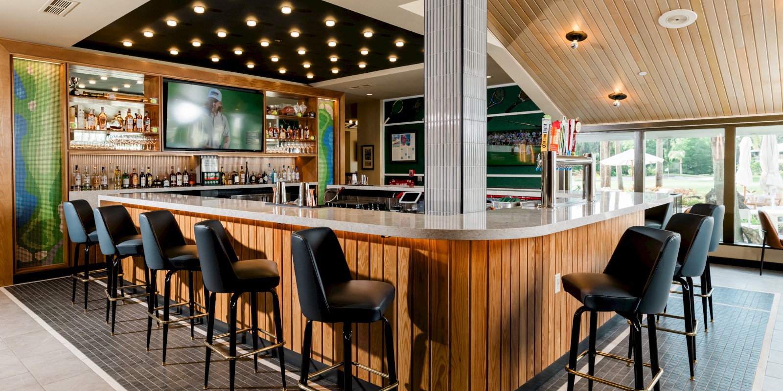 A modern indoor bar with black chairs, wooden accents, a large TV, and large windows overlooking a patio.