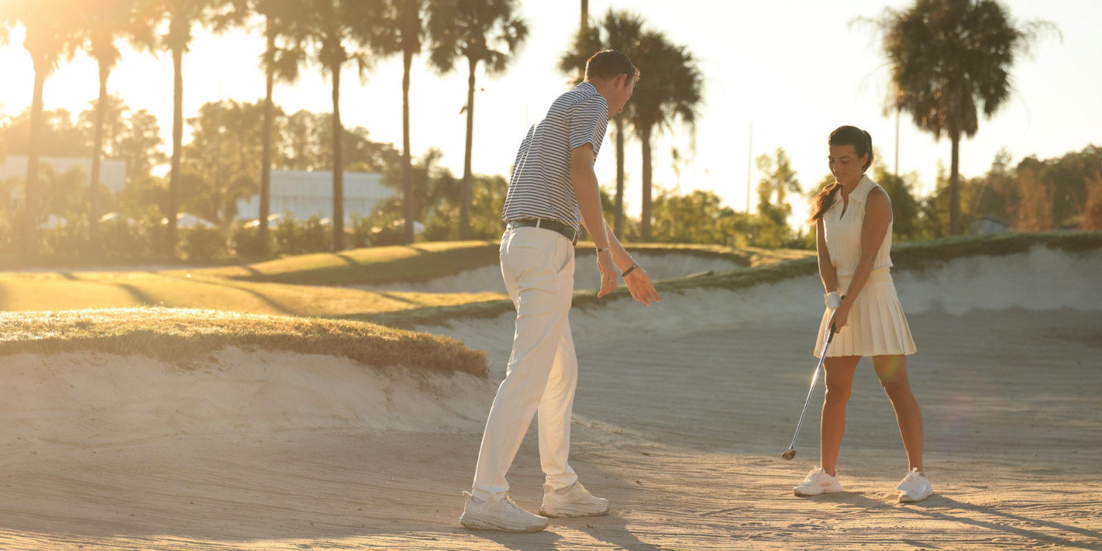 Two people playing golf on a sunny beach golf course, with palm trees in the background; a casual moment on the green, warm light.