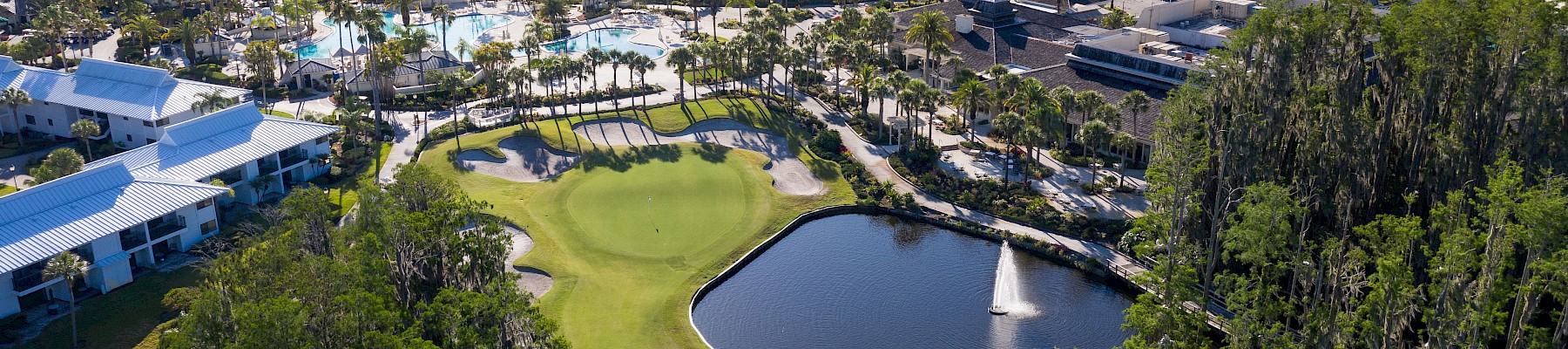 Aerial view of a golf course with a pond, nearby buildings, and surrounding greenery under a clear sky.