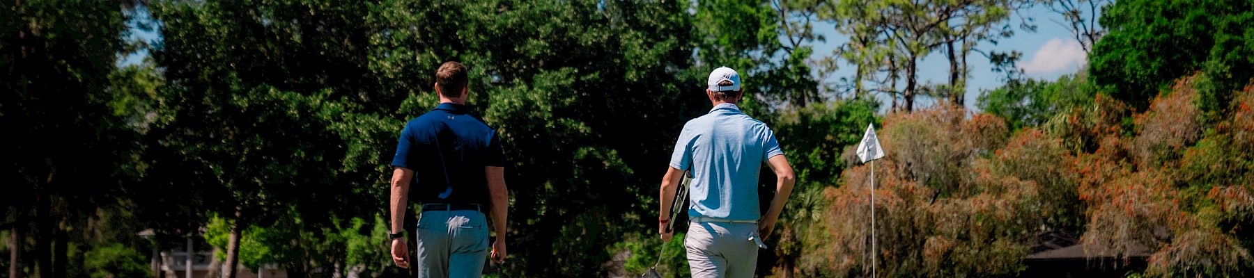 Two people walking on a golf course towards a flag, surrounded by trees and clear blue sky.