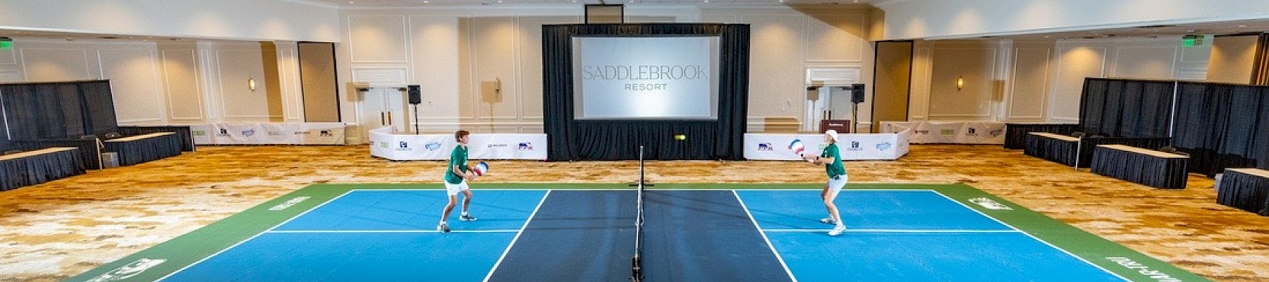 Two people are playing pickleball on a blue court set up indoors, surrounded by conference-style seating and a large screen.