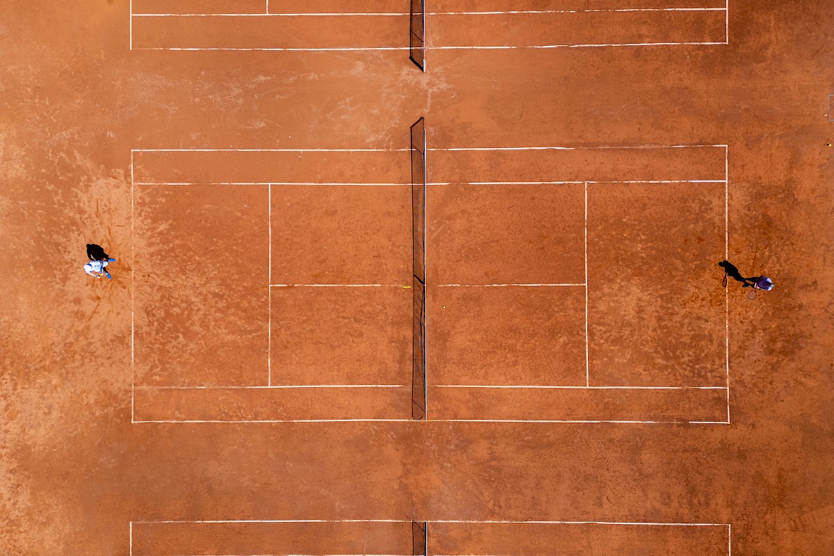 An aerial view of two people playing on a clay tennis court, with lines and net visible.