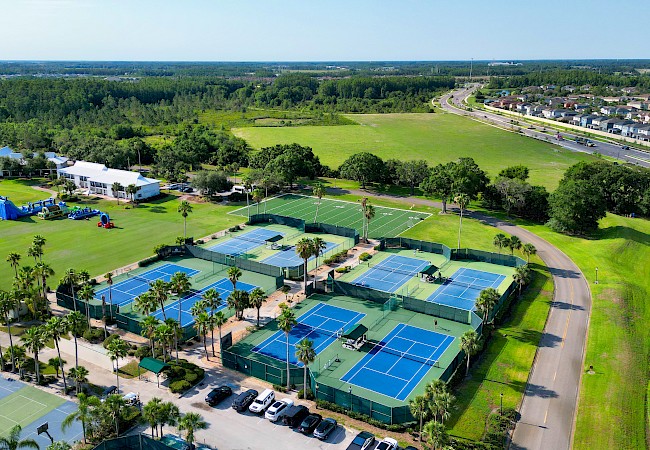 The image shows a sports complex with multiple tennis courts, surrounded by greenery and nearby roads and buildings.