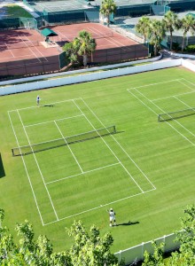 The image shows a grassy tennis court with two tennis players, surrounded by other courts and lush greenery.