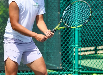 A person in tennis attire holds a racket, standing in front of a green chain-link fence on a tennis court.