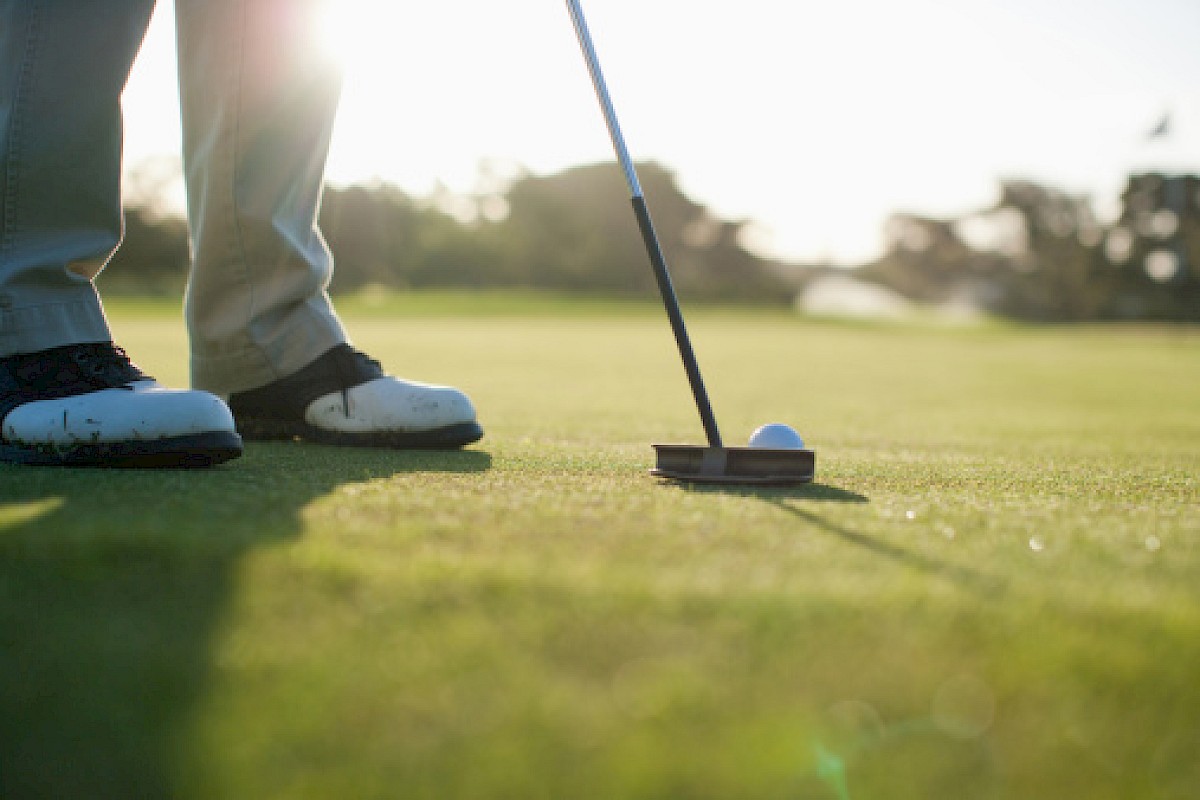 A person is on a golf course getting ready to putt a golf ball into the hole with a putter, under sunlight.