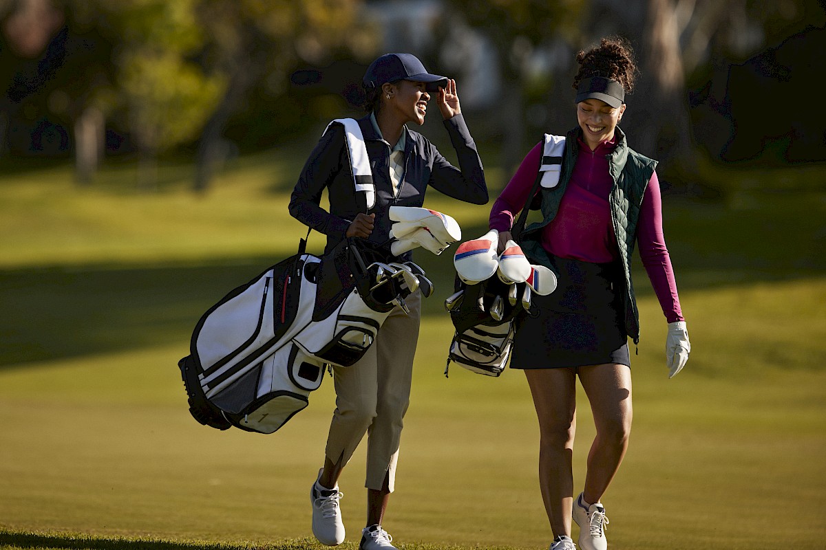 Two people are walking on a golf course, carrying golf bags and smiling, in a sunny outdoor setting.
